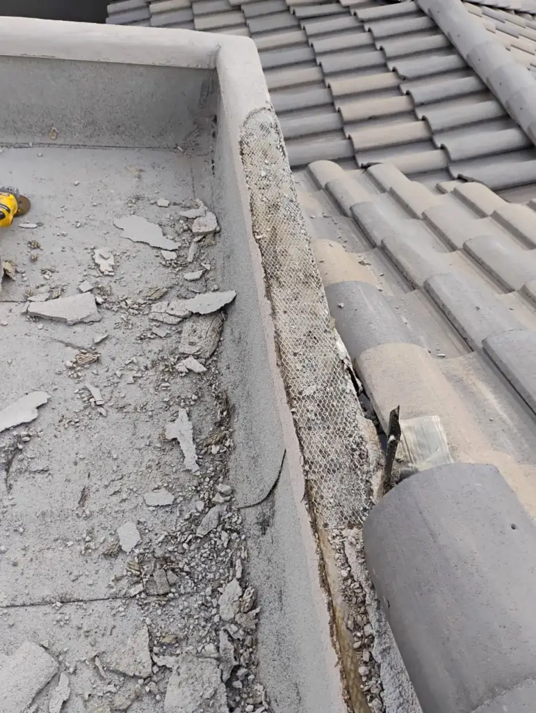 Top of a parapet wall with cracked, broken stucco next to roof tiles