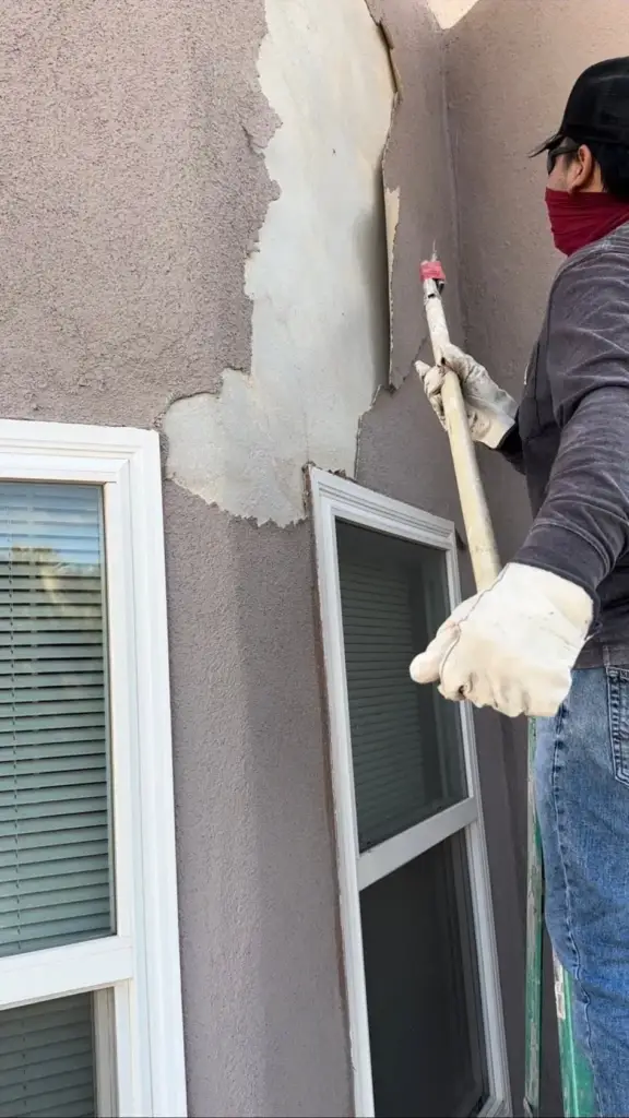 Plasterer knocking out loose stucco and filling a damaged area beside a window