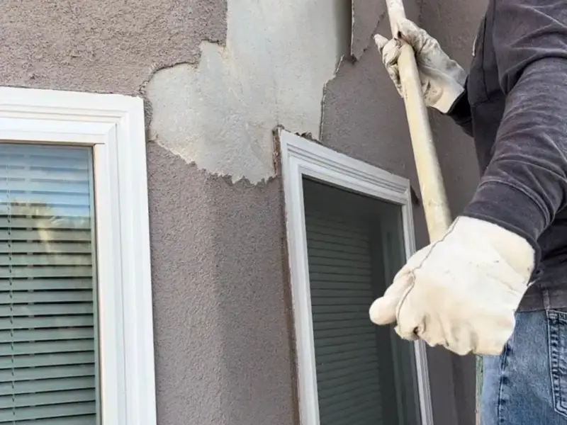 Plasterer knocking out loose stucco and filling a damaged area beside a window